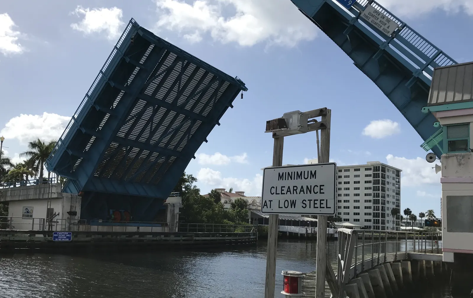 The blue steel leaves of a drawbridge raised against a partly cloudy sky over dark water, with a "MINIMUM CLEARANCE AT LOW STEEL" sign in the foreground and a white high-rise building behind.