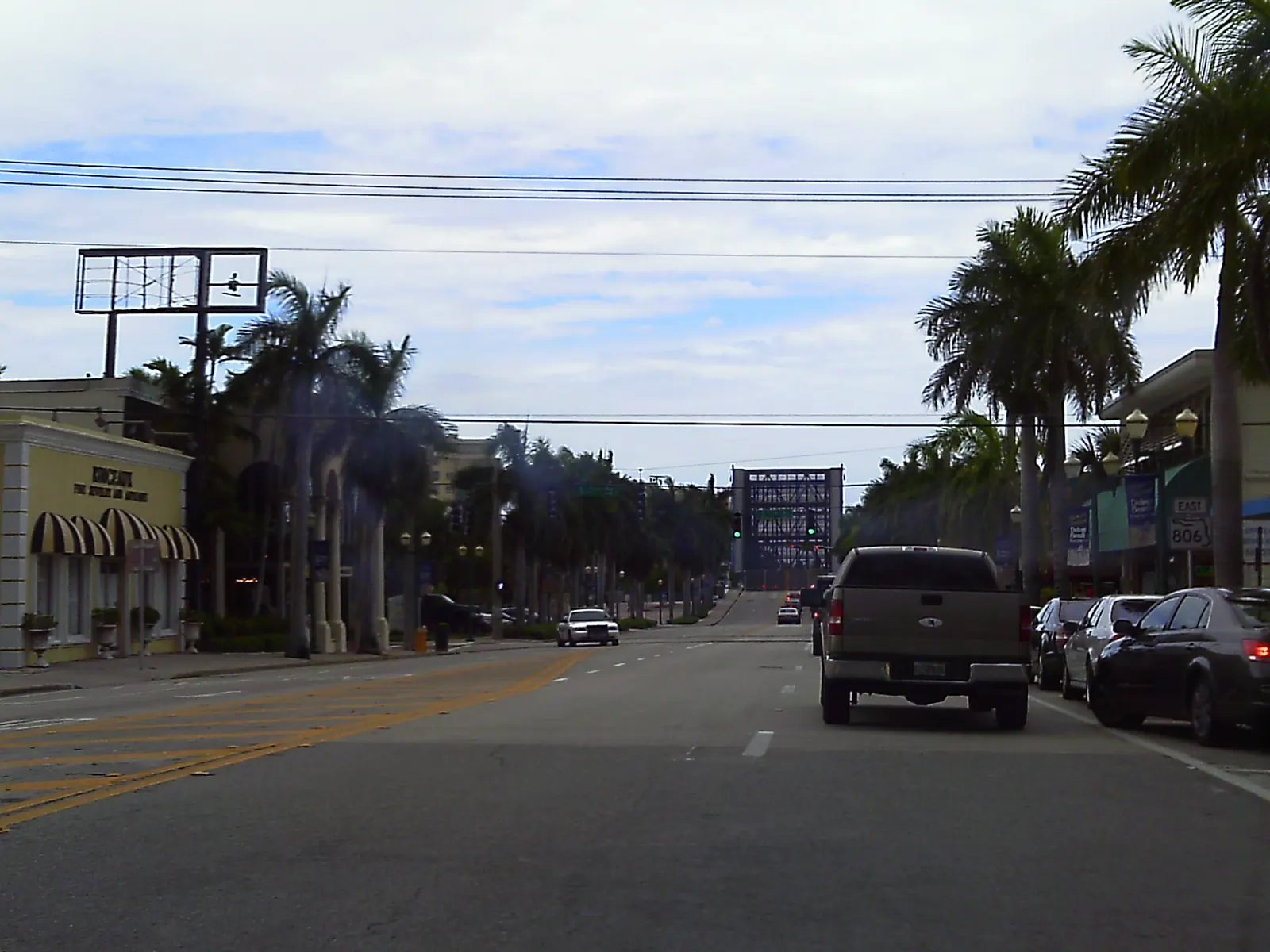 Daytime view east down a multi-lane avenue: a dark pickup truck and parked cars on the right, tall palms along both sides, low shop buildings with awnings at left, overhead utility wires and a raised drawbridge span visible at the far end of the street.