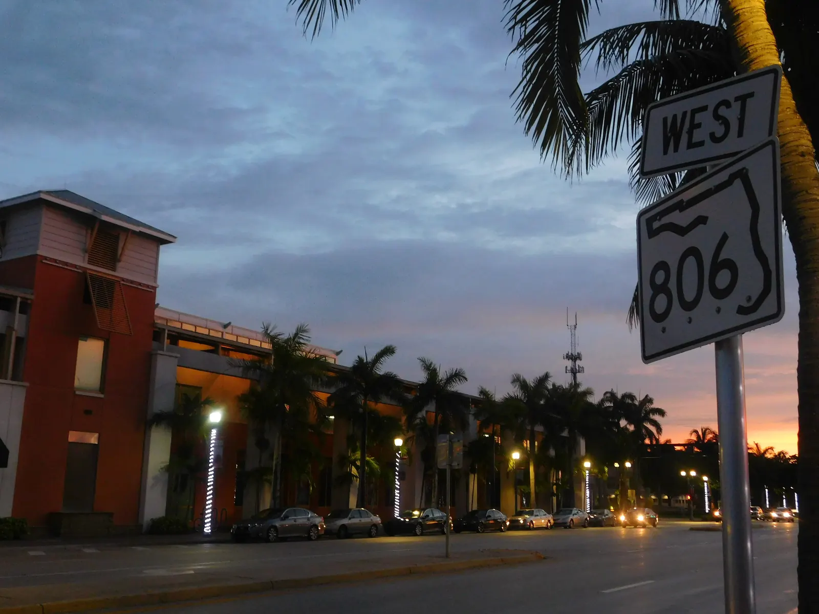 Dusk scene with a "WEST 806" state-road shield on a pole at right, palm trees silhouetted against a blue-and-pink sky, a lit row of street lamps and a multi-storey building with illuminated columns along the left side of the road.