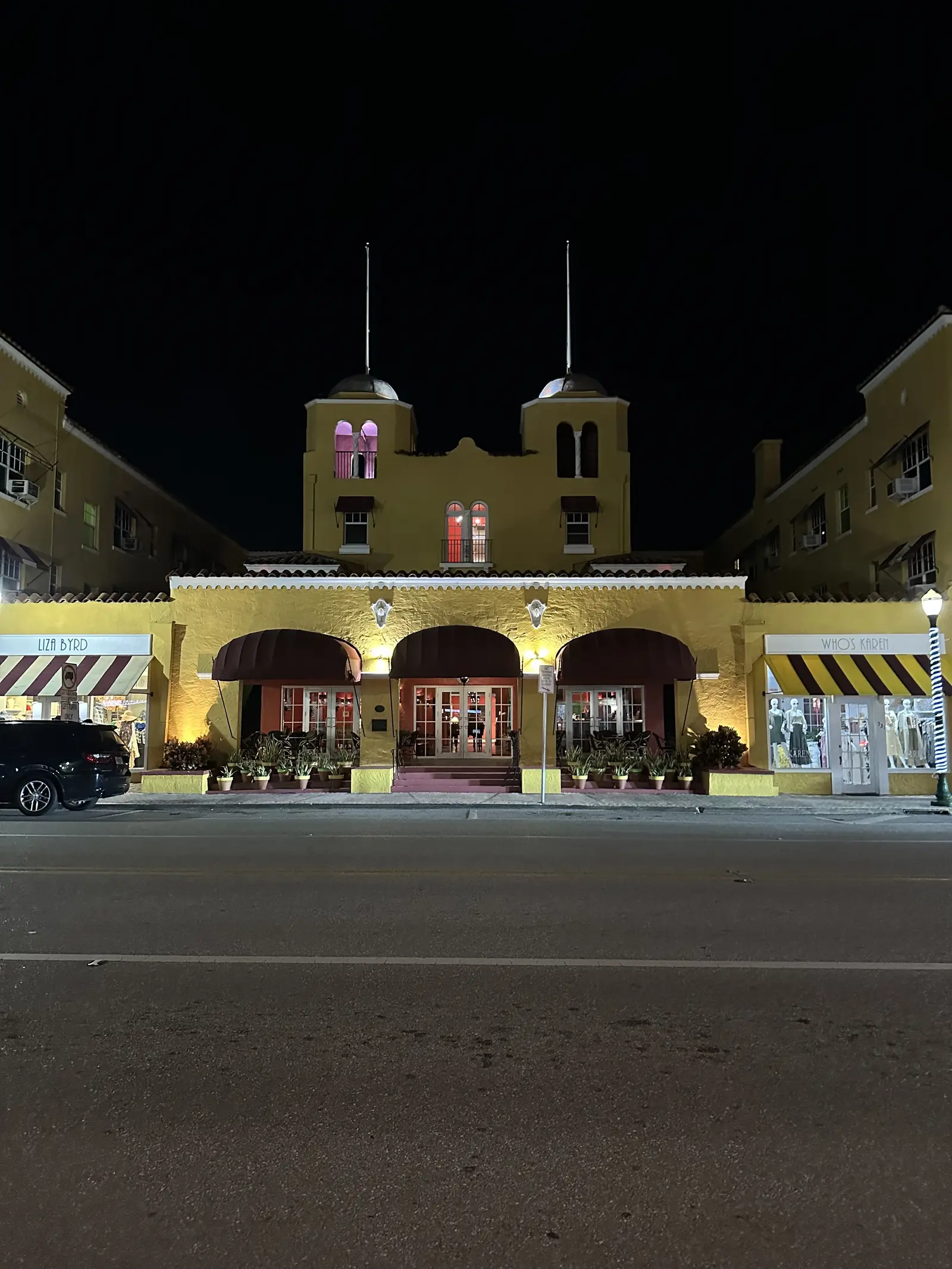 Night view of a yellow Mediterranean-revival hotel with twin towers and flagpoles, a lit arched entrance with striped awnings, and an empty street in the foreground.