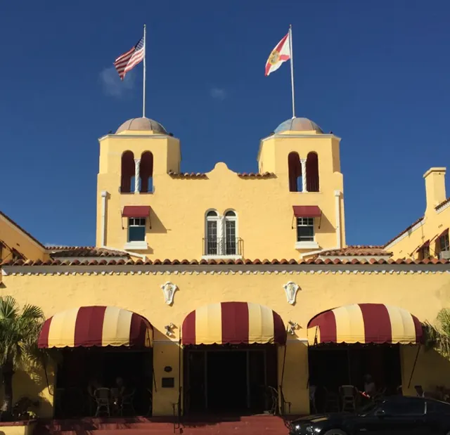Close view of the yellow twin-towered facade of the Colony Hotel with American and Florida flags flying on top, red-and-yellow striped awnings over the entrance, against a deep blue sky.
