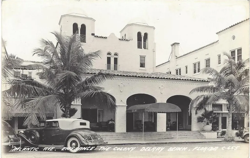 Black-and-white real-photo postcard of an arcaded Spanish-style hotel entrance with two bell-tower turrets, palms and a 1930s car parked in front; the caption along the bottom reads "Atlantic Ave. Entrance, The Colony, Delray Beach, Florida".
