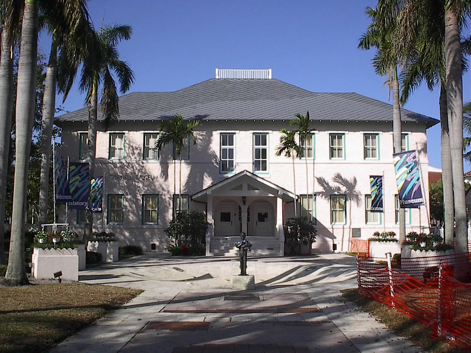 A two-storey white building with a grey hip roof and a small central porch, flanked by tall palms and banners, with a paved forecourt and an orange safety fence on the right.