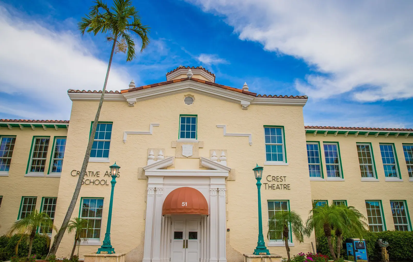 Front of a cream two-storey school building with green window trim, a central arched doorway under an orange awning, palms and green lamp posts, and lettering for "Creative Arts School" and "Crest Theatre" on the facade, under a blue sky with clouds.