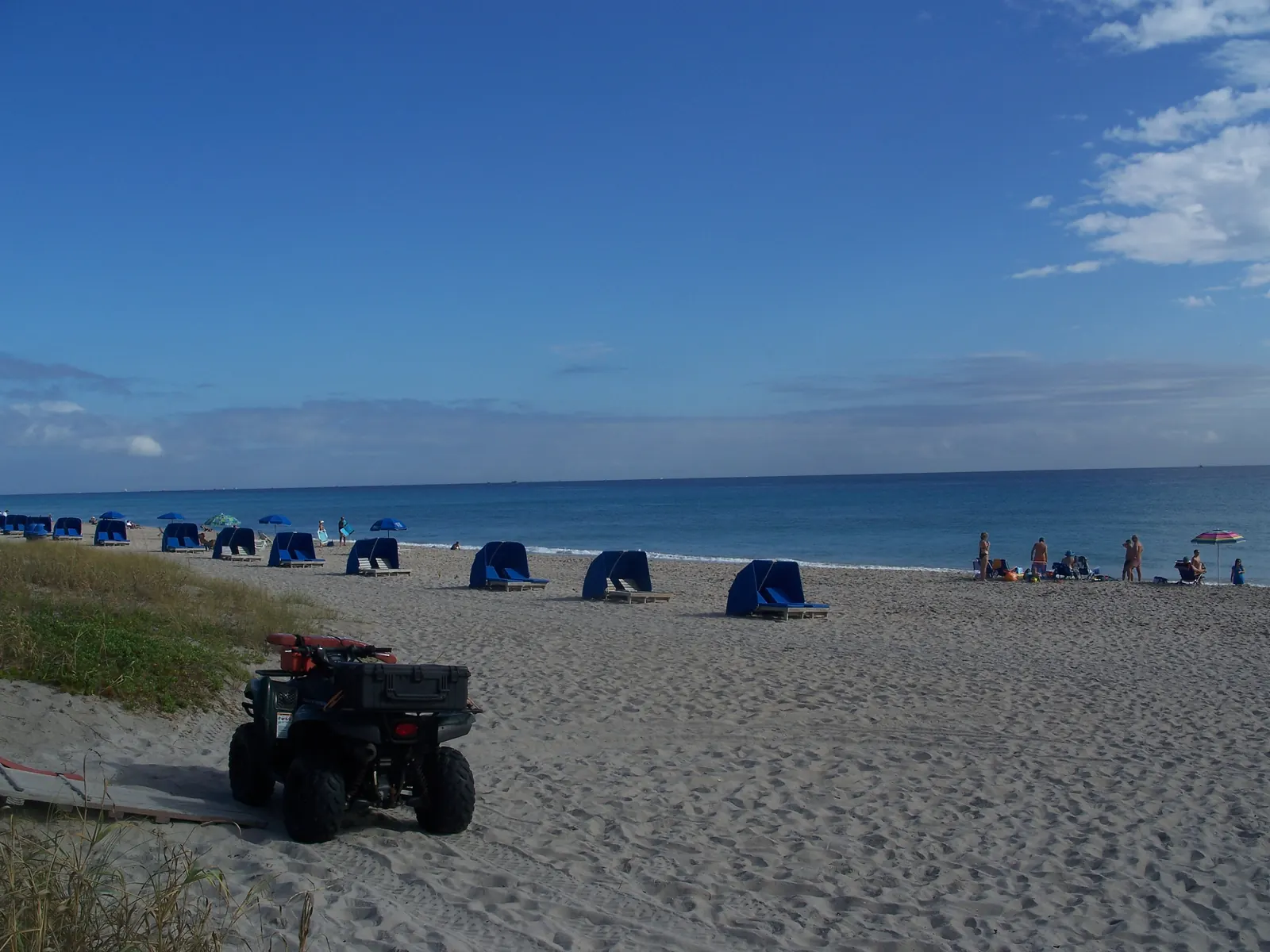 A wide sandy beach under a blue sky with rows of blue cabanas and umbrellas along the waterline, a few beachgoers at the surf and a red-and-black ATV parked on the sand in the foreground beside dune grass.