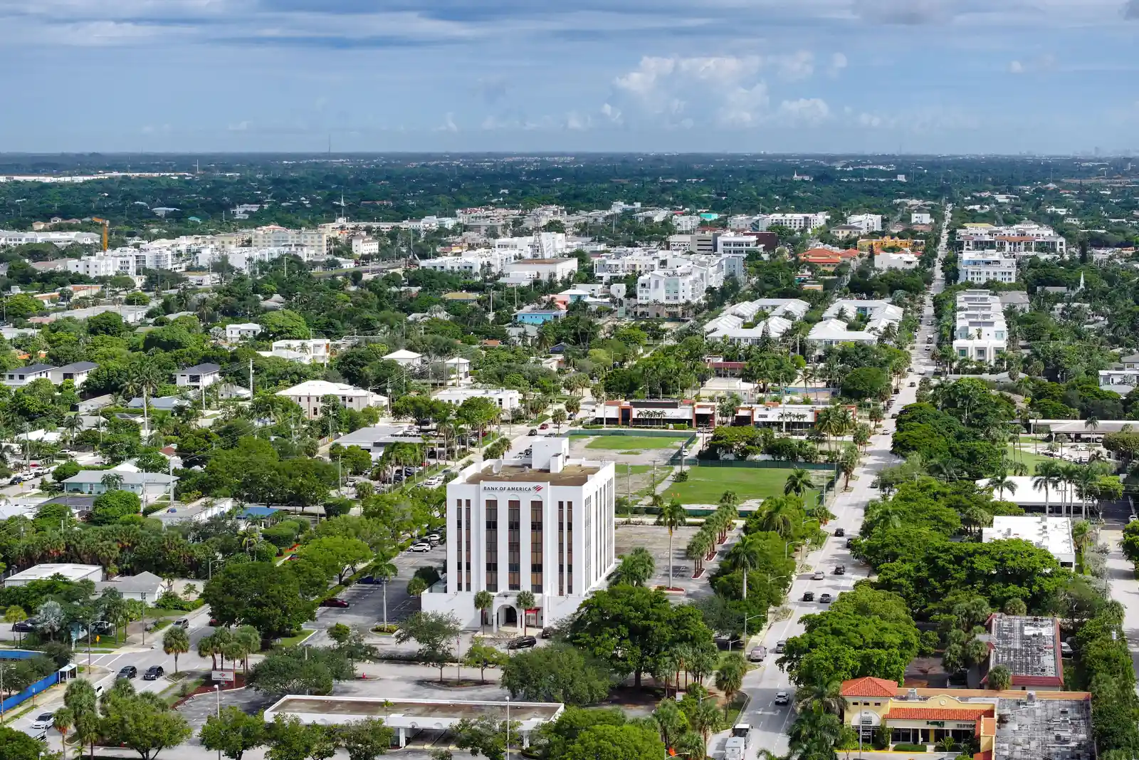 Aerial view over a low-rise town of white and pastel buildings and dense green tree canopy, with a white multi-storey office tower in the centre foreground and straight streets running toward the horizon.