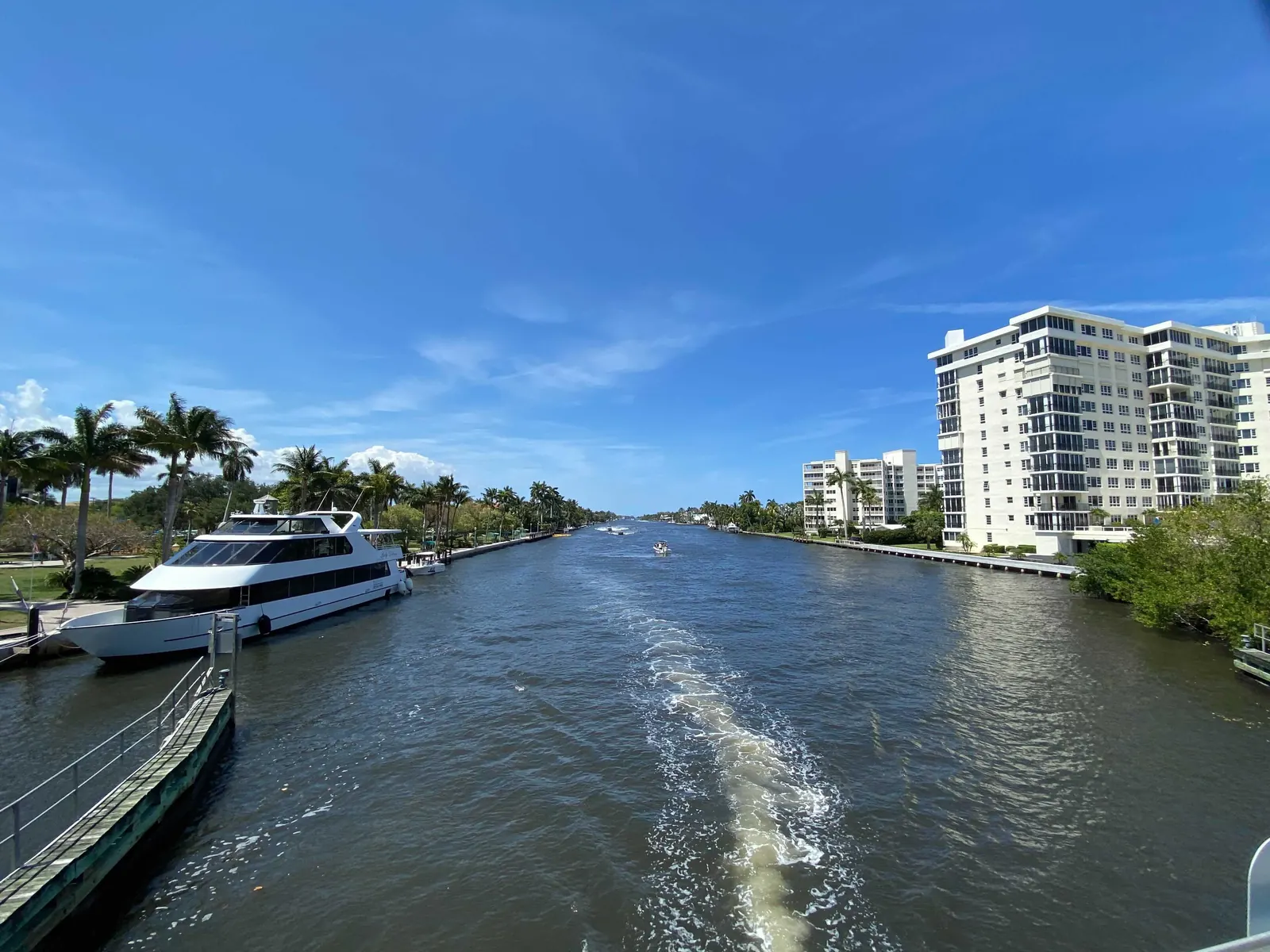 A wide waterway stretching into the distance under a blue sky, with a white yacht moored at a dock on the left, a boat wake in the middle and white high-rise condominium buildings on the right bank.