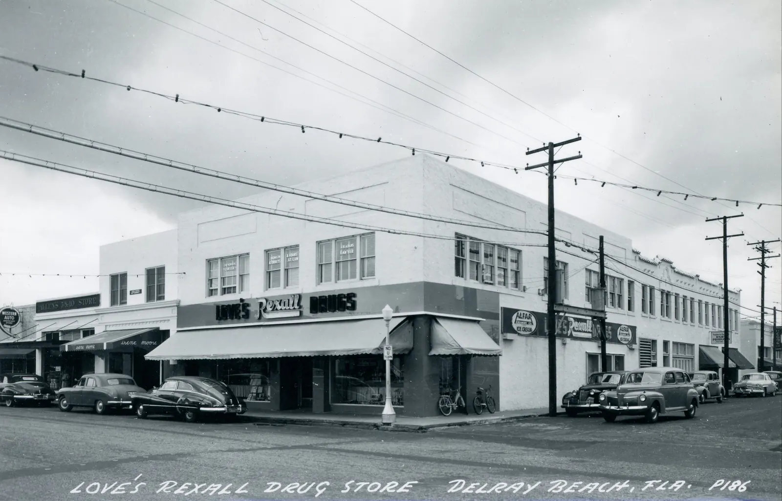 Black-and-white photo postcard of a two-storey corner commercial building with a "Love's Rexall Drugs" sign and awnings, 1940s cars parked along the curb and overhead wires; caption reads "Love's Rexall Drug Store, Delray Beach, Fla."