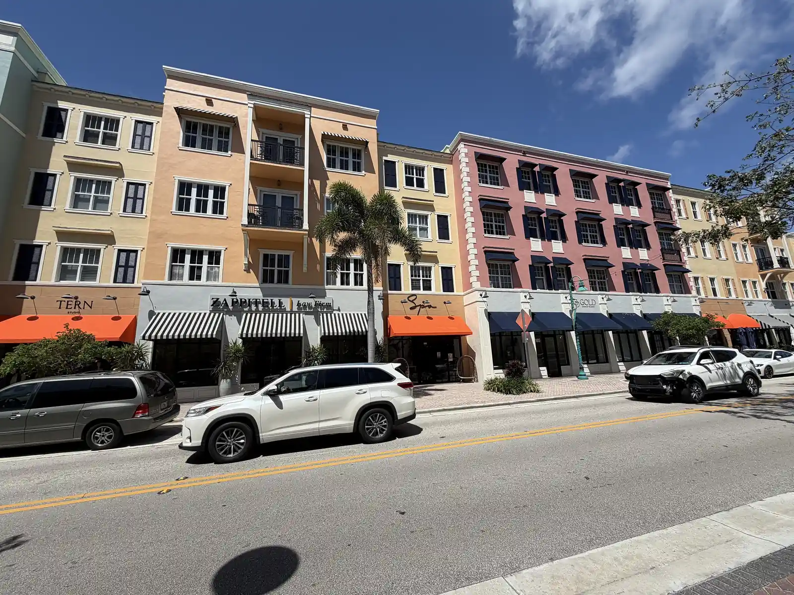 Sunny street view of a continuous row of three- and four-storey stucco buildings in peach, pink and cream, with orange and striped awnings over ground-floor restaurants and shops; SUVs are parked and driving along the street in front.