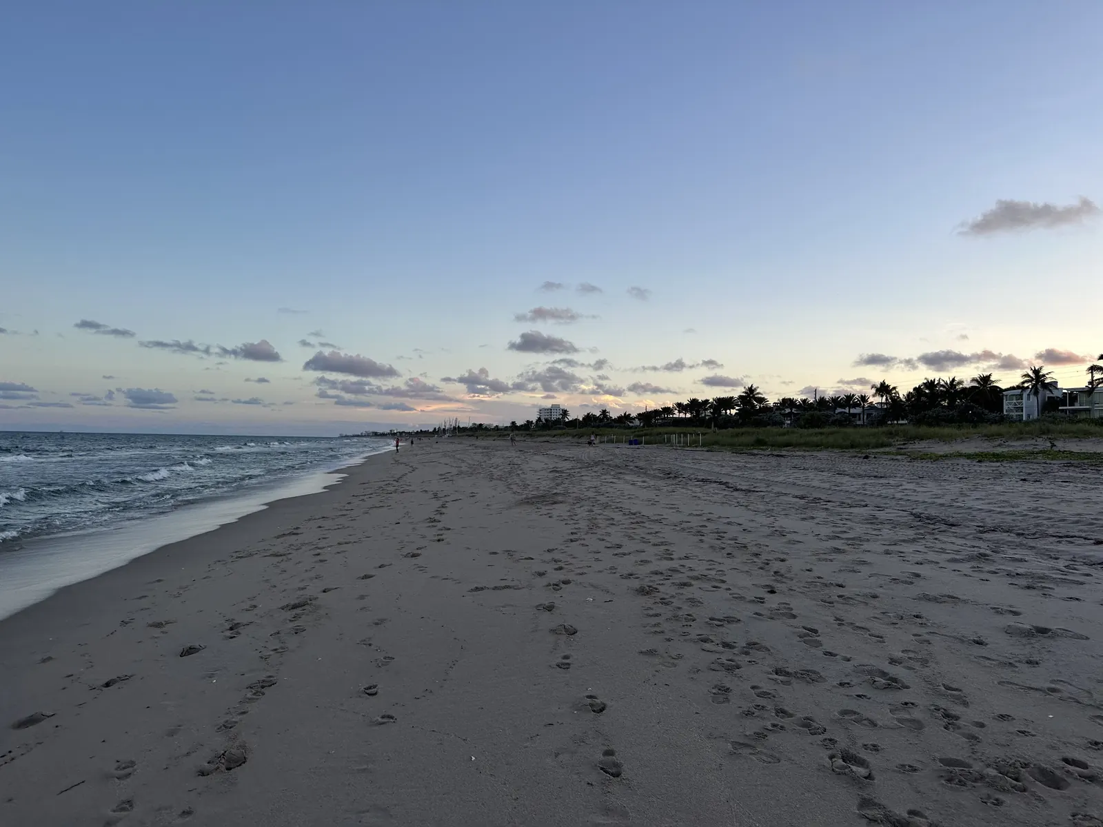 A long, empty beach with footprints in the sand stretching south under a pale evening sky, gentle surf on the left and low beachfront trees and houses on the horizon.