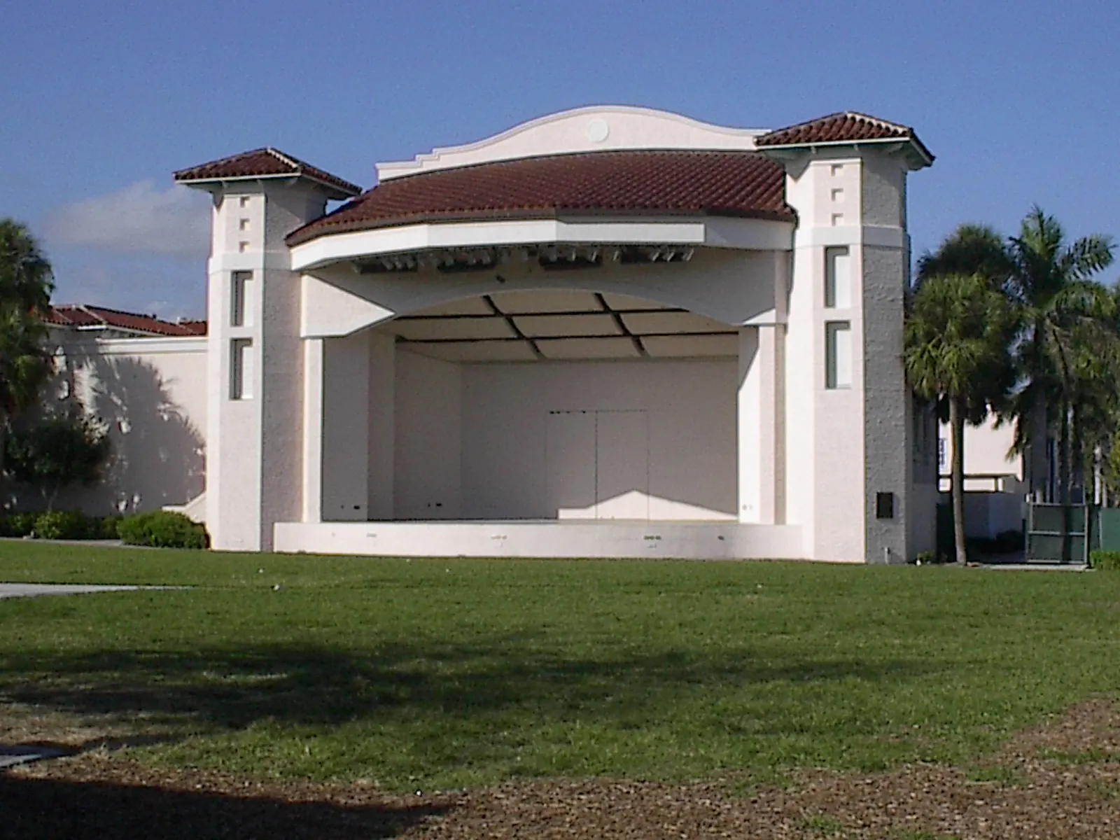 A white stucco open-air stage with a red tile roof and square side towers, facing an empty lawn under a blue sky.