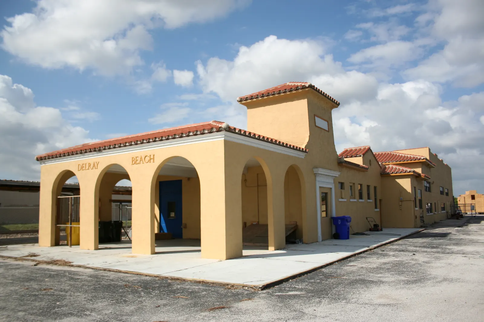 A yellow stucco railway station with a red tile roof, a square tower and an open arched loggia with "DELRAY BEACH" lettering, seen across an empty paved lot under a cloudy sky.