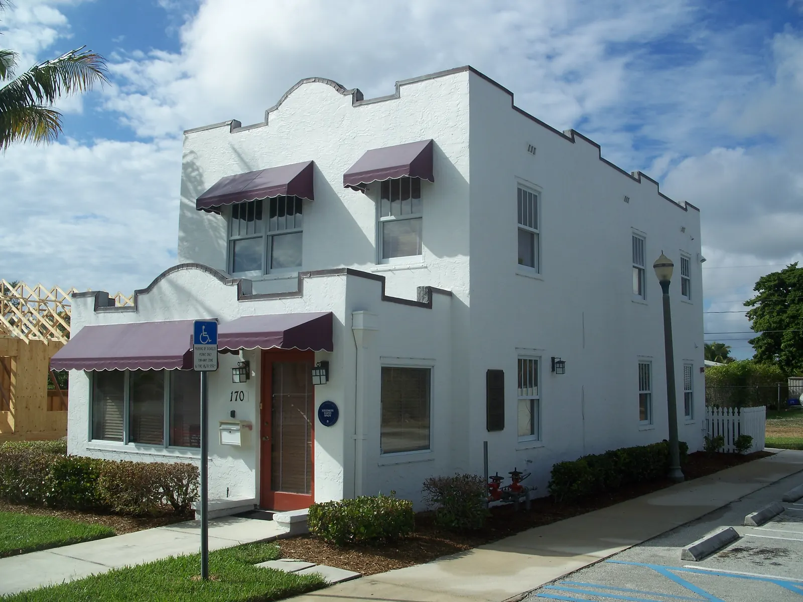 A white two-storey Mission-style house with a curved parapet, maroon fabric awnings over the windows, a red front door with the number 170, a lamp post and a disabled-parking sign, under a cloudy blue sky.